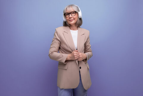 middle-aged woman in a stylish jacket listens to music in large headphones on a bright background with copy space.